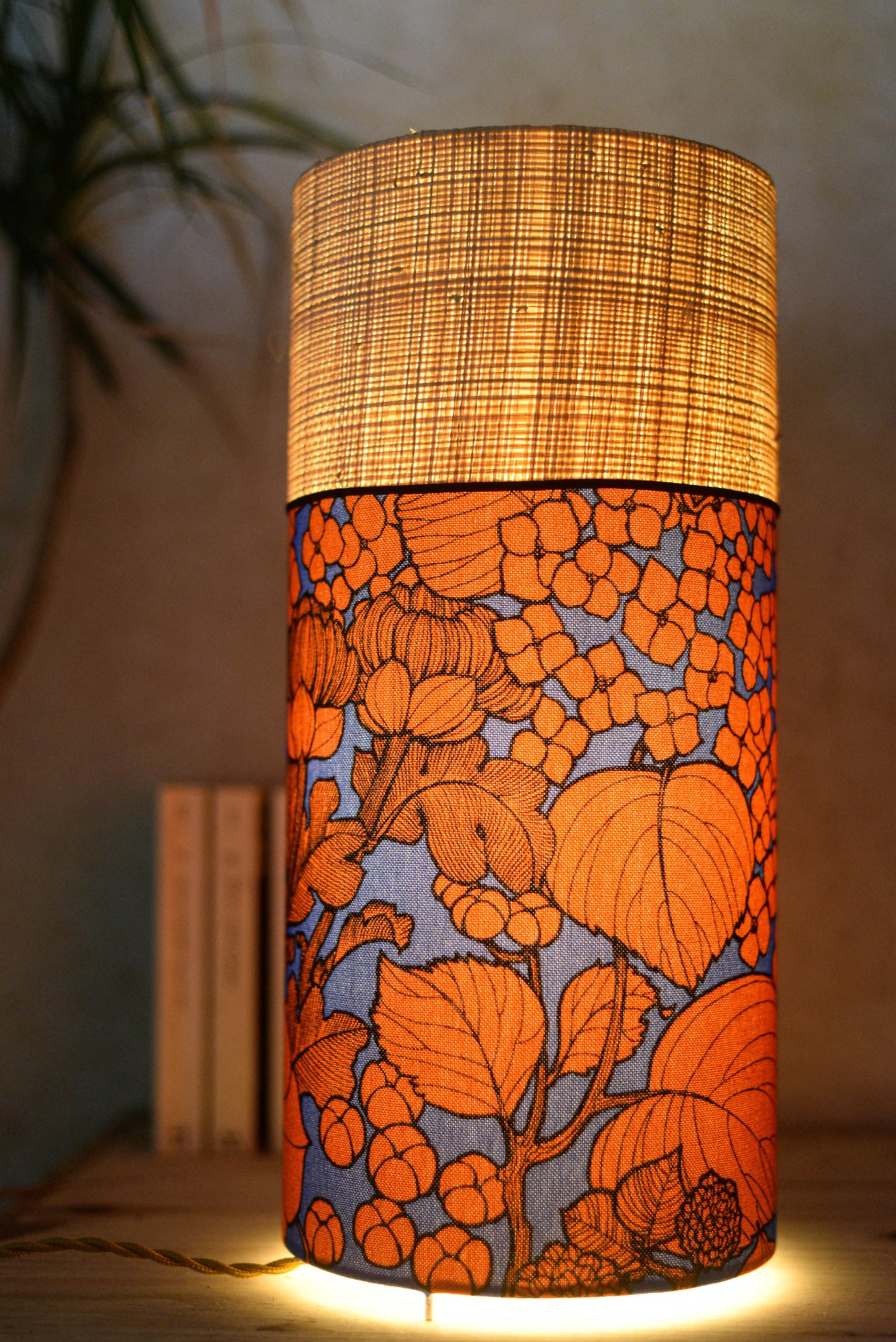 Lampe tube cynlindrique bi-matière en raphia naturel et tissu à motif de branches et fleurs orange sur fond bleu. Tissu de la collection Thevenon