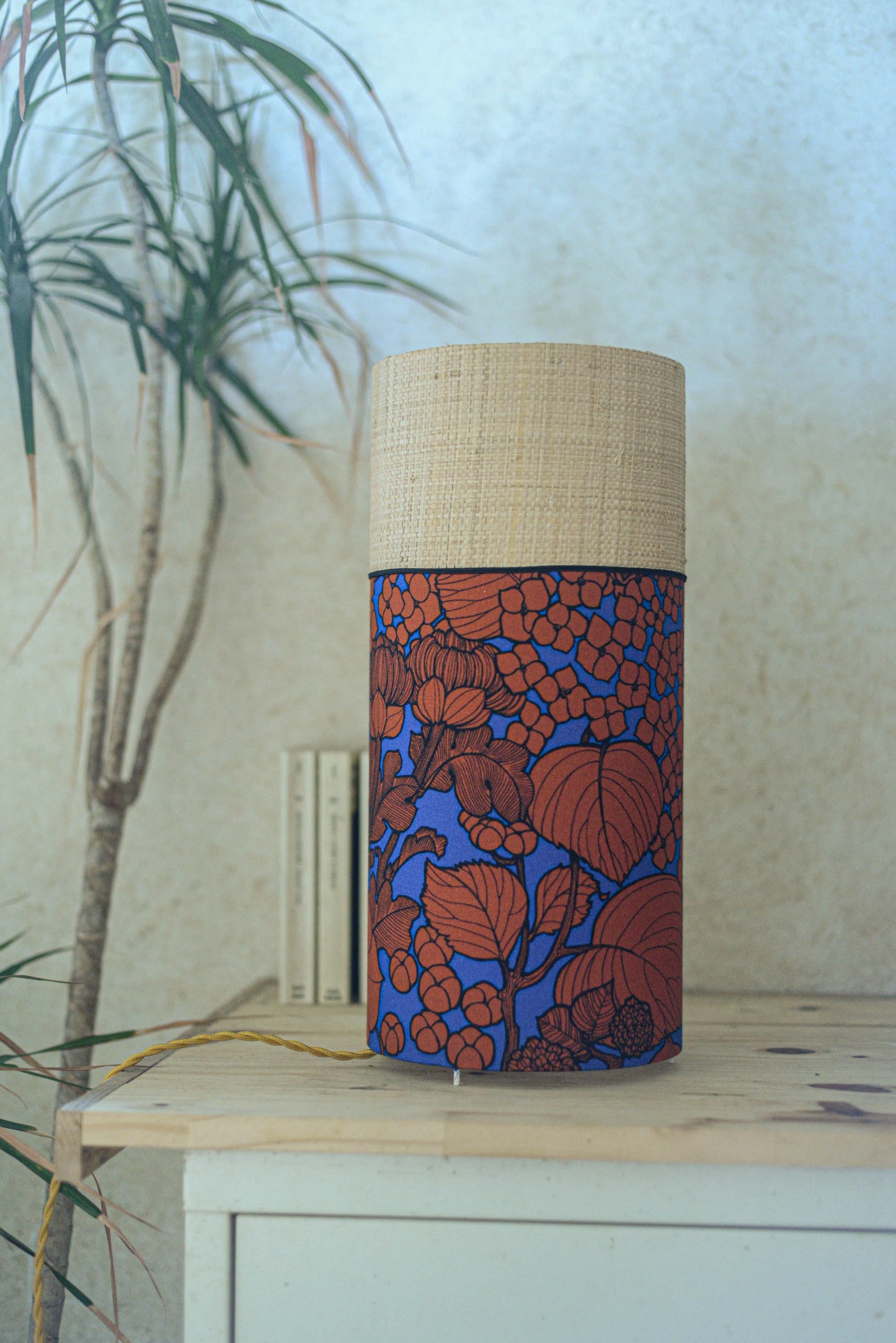 Lampe tube cynlindrique bi-matière en raphia naturel et tissu à motif de branches et fleurs orange sur fond bleu. Tissu de la collection Thevenon