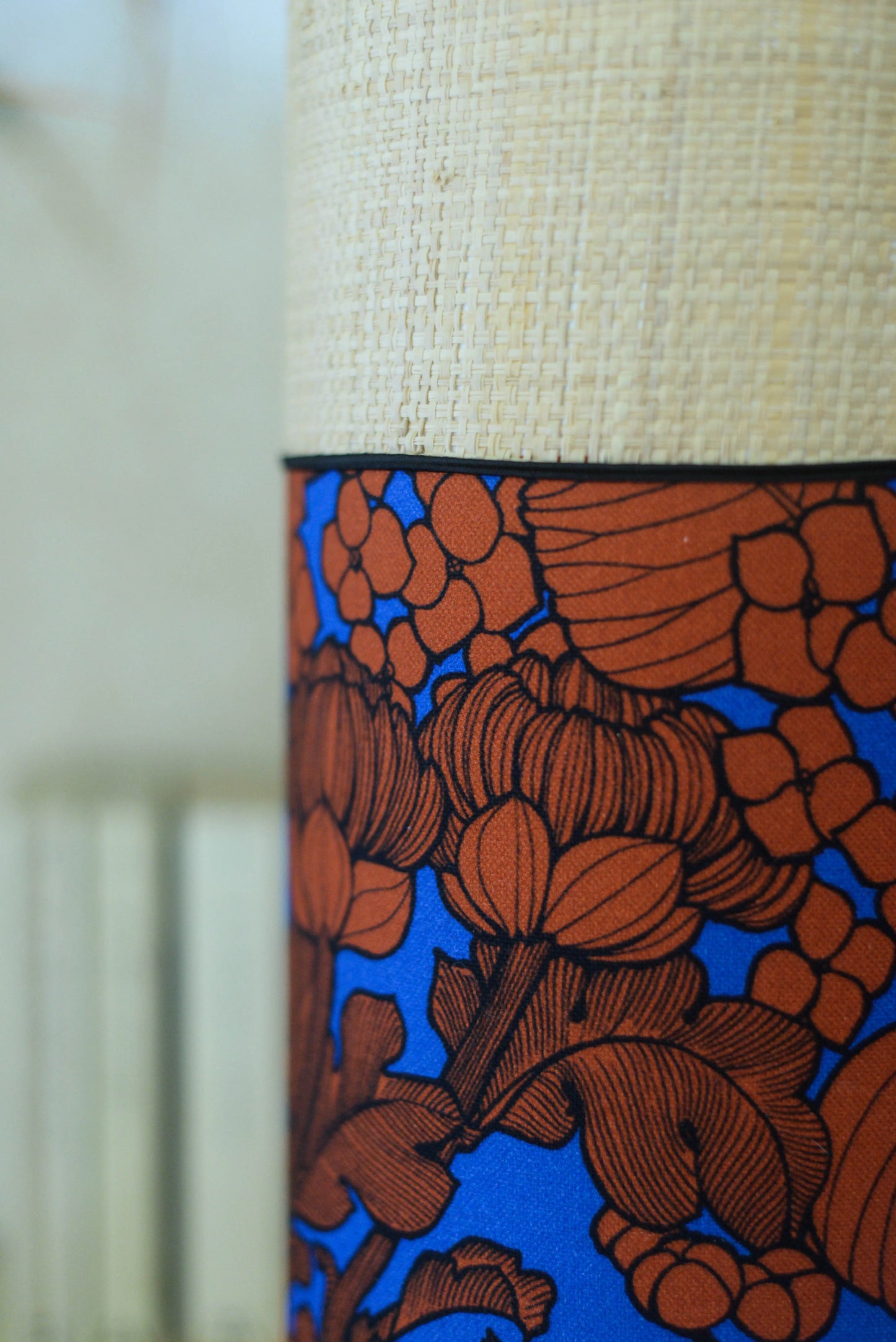 Lampe tube cynlindrique bi-matière en raphia naturel et tissu à motif de branches et fleurs orange sur fond bleu. Tissu de la collection Thevenon