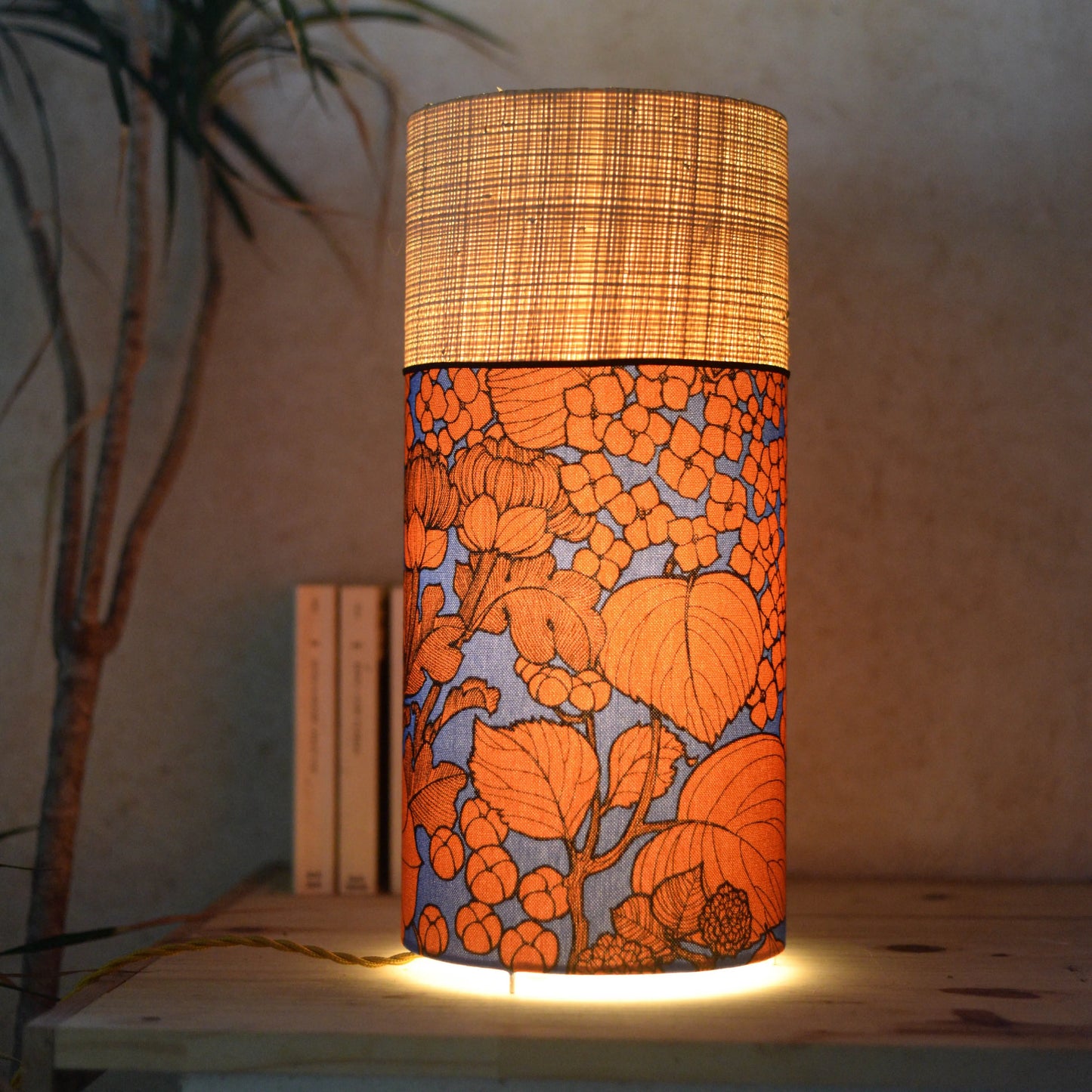 Lampe tube cynlindrique bi-matière en raphia naturel et tissu à motif de branches et fleurs oranges sur fond bleu. Tissu de la collection Thevenon