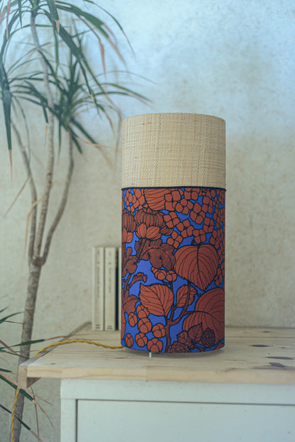 Lampe tube cynlindrique bi-matière en raphia naturel et tissu à motif de branches et fleurs orange sur fond bleu. Tissu de la collection Thevenon