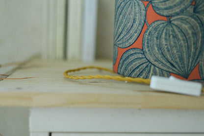 Vue du cordon torsadé doré de la lampe tube cynlindrique bi-matière en raphia naturel et tissu à motif de physalis bleu écru sur fond orange. Tissu de la collection Thevenon