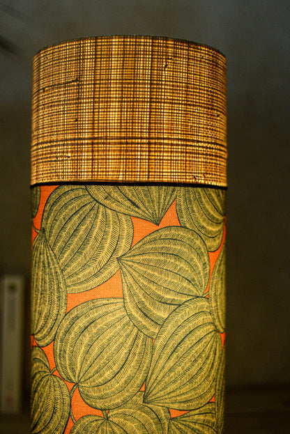 Lampe tube cynlindrique bi-matière en raphia naturel et tissu à motif de physalis bleu écru sur fond orange. Tissu de la collection Thevenon